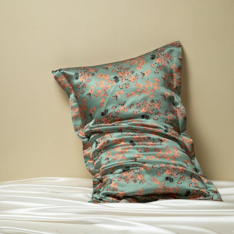 Sage green silk pillowcase printed with coral and orange blossoms, leaning on a cream wall above white silk