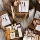 Open wooden gift box with soaps and skincare gifts beside a closed box with a caramel ribbon and orchid flower