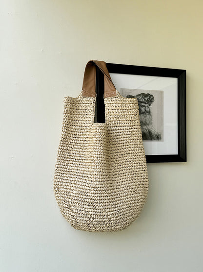 Beige paper straw woven shoulder bag hanging by its tan straps on a wall beside a framed print
