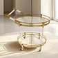 Round gold finish serving cart with glass tiers and wheels on a rug in a bright room