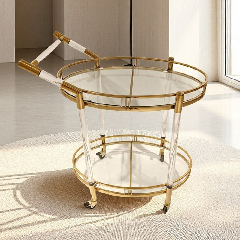 Round gold finish serving cart with glass tiers and wheels on a rug in a bright room