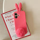 Deep pink silicone iPhone case with 3D rabbit ears and a plush pom pom tail on a red book