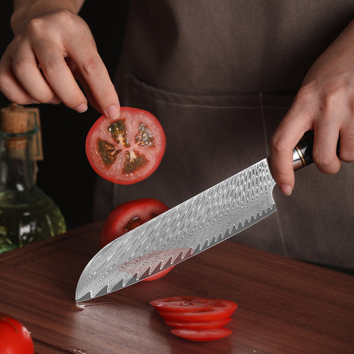 Damascus pattern santoku knife slicing a tomato on a wooden board with whole tomatoes and an oil bottle behind