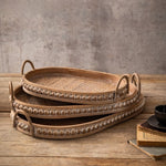 Three stacked oval rattan trays with studded rims and arched handles on a wooden table with a dark cup