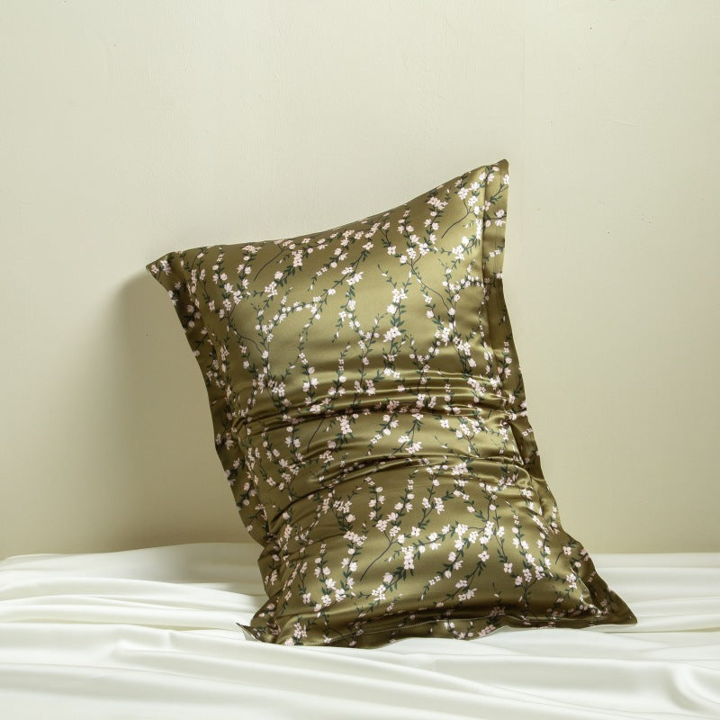 Olive green silk pillowcase printed with white blossom vines, leaning on a cream wall above white silk