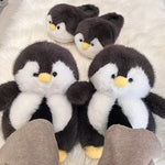 Dark gray plush penguin slippers with yellow beaks and black eyes, two pairs on a white fluffy rug, one pair worn