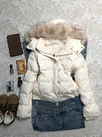Cream white hooded quilted jacket with a furry hood trim laid flat on lace beside jeans and accessories