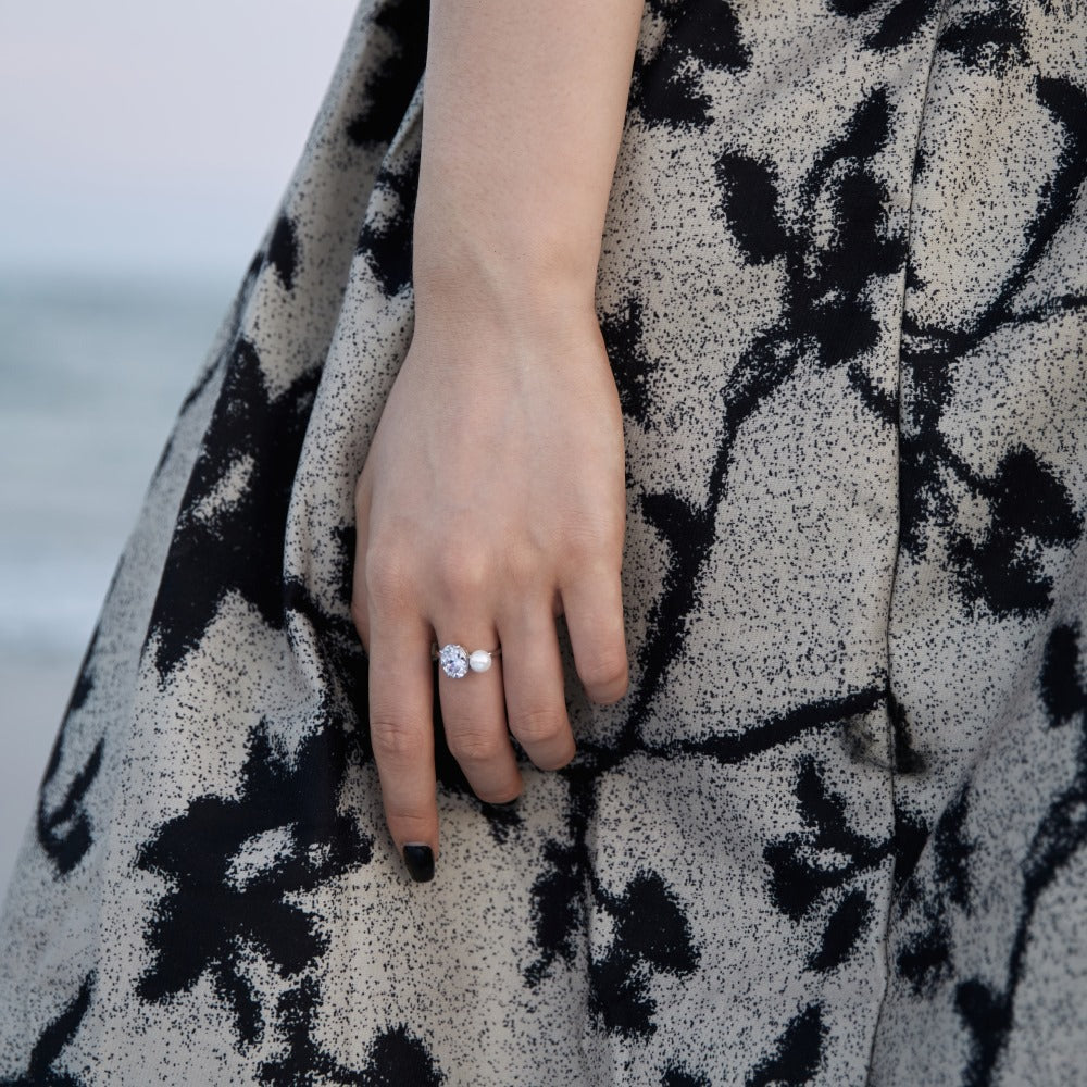 Hand at the shore wearing the platinum toned open ring, oval stone and imitation pearl in daylight