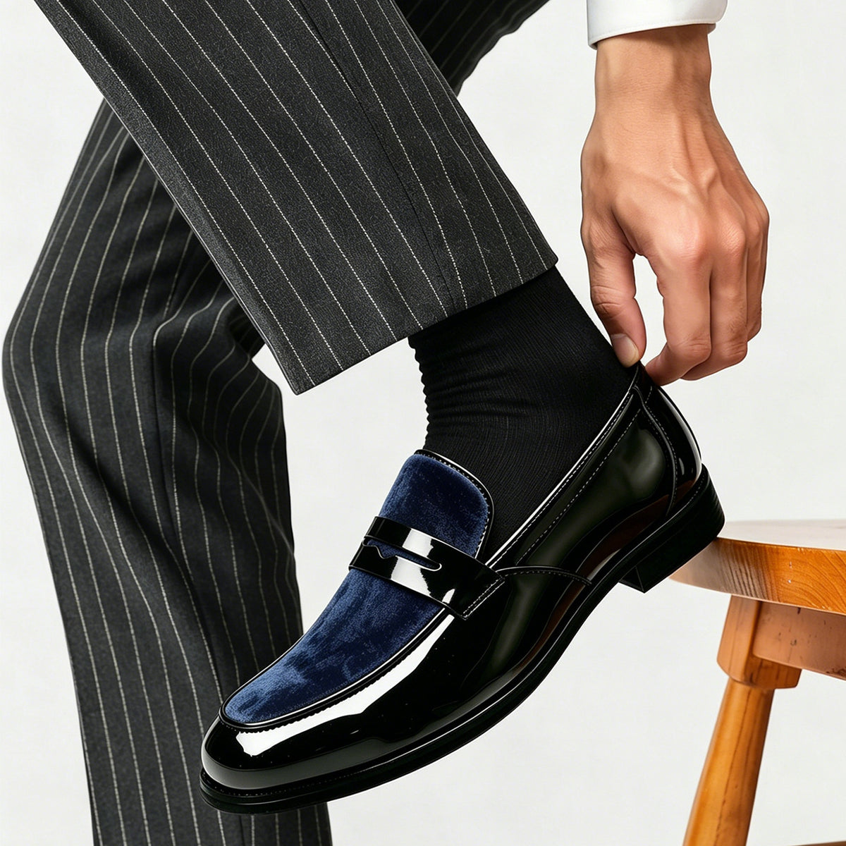 Man in pinstripe trousers putting on a glossy black loafer with a navy velvet vamp and a penny strap