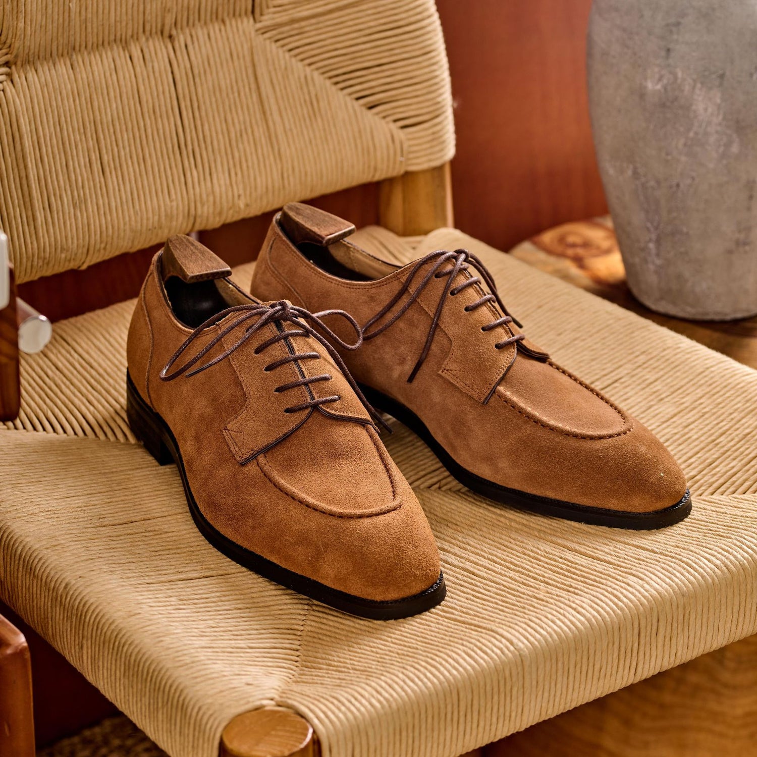 Brown suede apron toe derbys angled on a woven chair seat, low stacked heel and dark rubber outsole edge