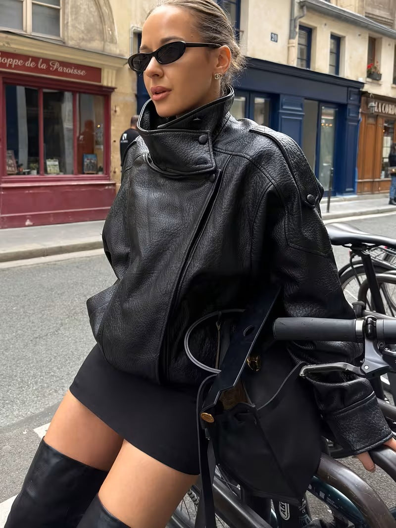 Woman in sunglasses by a shopfront in a black oversized PU leather moto jacket with a stand collar