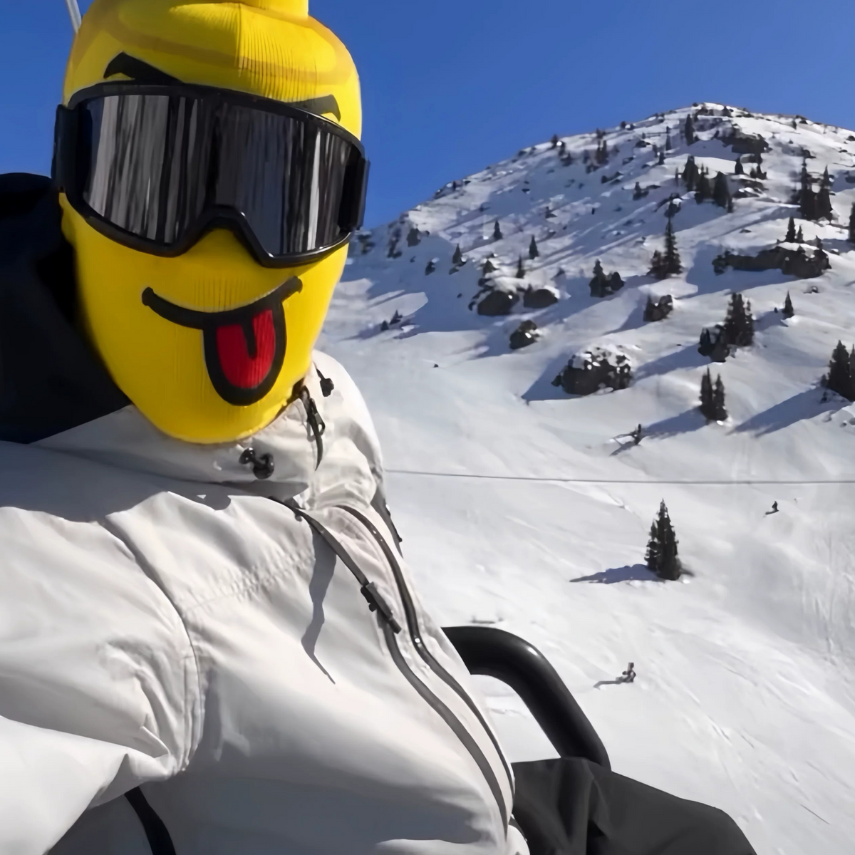 Yellow block head printed face cover with tongue out and ski goggles beside a snowy mountain slope