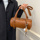 Hand holding a cognac brown cylinder shoulder bag with embossed lettering, buckle strap and baseball charm