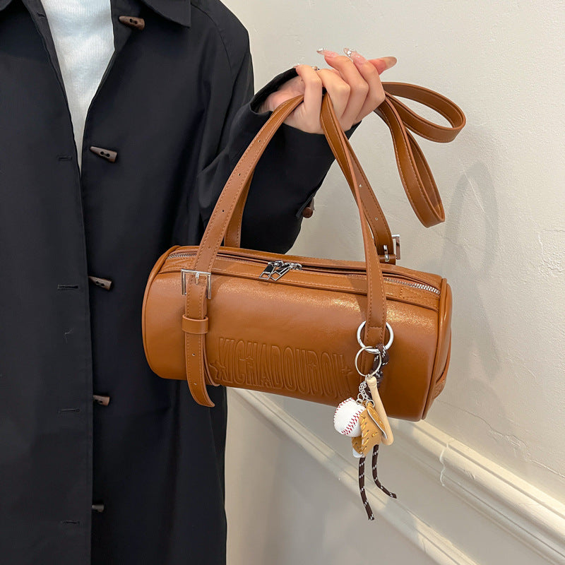 Hand holding a cognac brown cylinder shoulder bag with embossed lettering, buckle strap and baseball charm