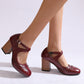 Wine red brogue Mary Jane pumps with a buckled cross strap and block heels, worn on a light studio floor