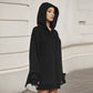 Woman in a black oversized hoodie seen from the side on a street, with the pointed wing edge of one sleeve hanging at the hip