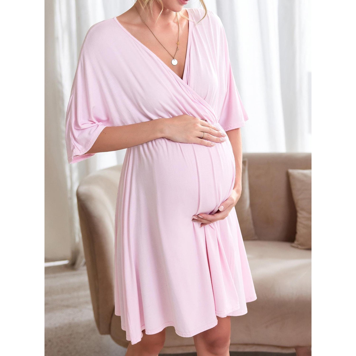 Pregnant model in a pastel maternity wrap dress standing in a sunlit living room