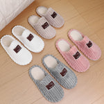 Pairs of ribbed plush slippers in white, pink, khaki and gray lying on a wooden floor beside a bed