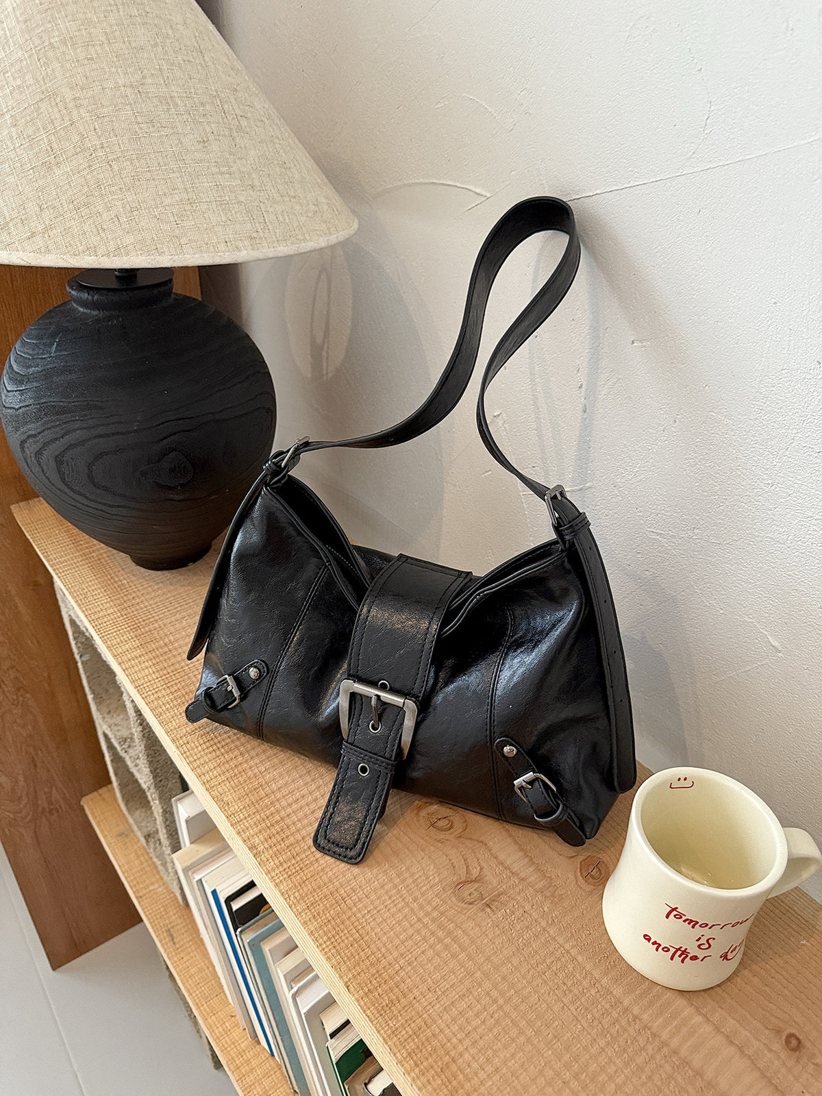 Black soft vegan leather hobo bag with front buckle strap and wide shoulder strap on a wooden shelf