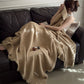 Woman holding the edge of a tan cotton gauze throw to her face on a dark leather sofa, fringe hanging along the edge
