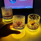 Hand cupping one of three lit cut glass whiskey tumblers on a dark desk