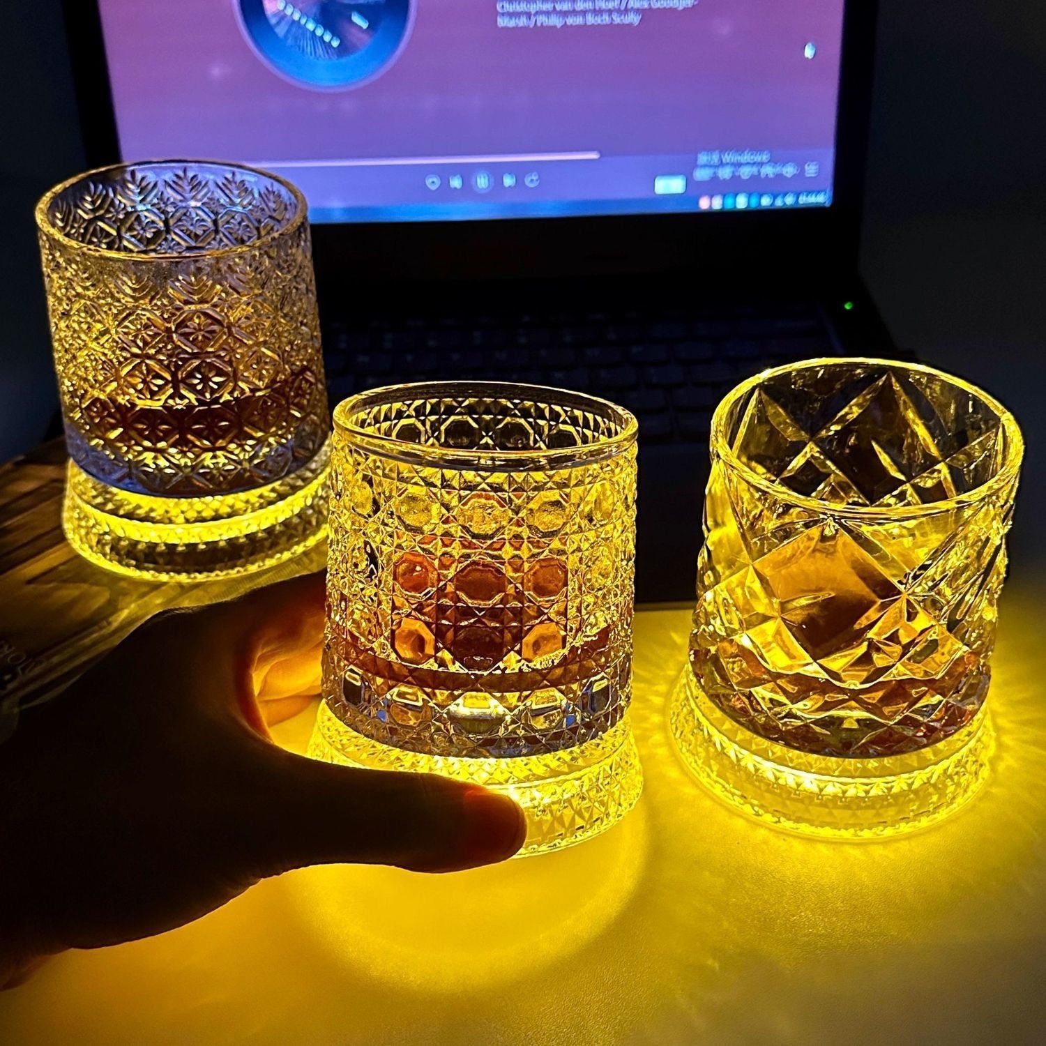 Hand cupping one of three lit cut glass whiskey tumblers on a dark desk