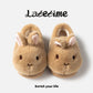 Pair of brown bunny plush slippers seen from above on a light panel with script lettering and a slogan, view 1 of 5