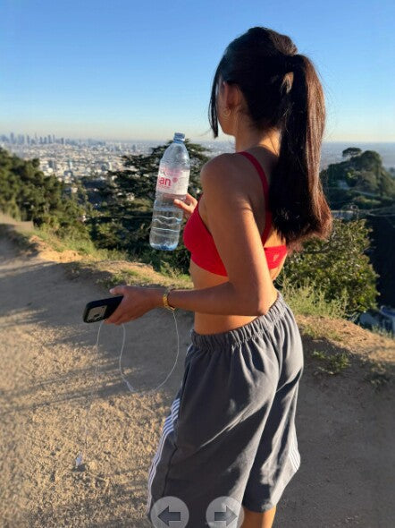 Woman in gray striped pants and a red top seen from behind on a hillside, holding a water bottle