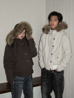 Couple wearing short hooded jackets with fur trimmed hoods, a dark brown one and an off white one, against a white wall