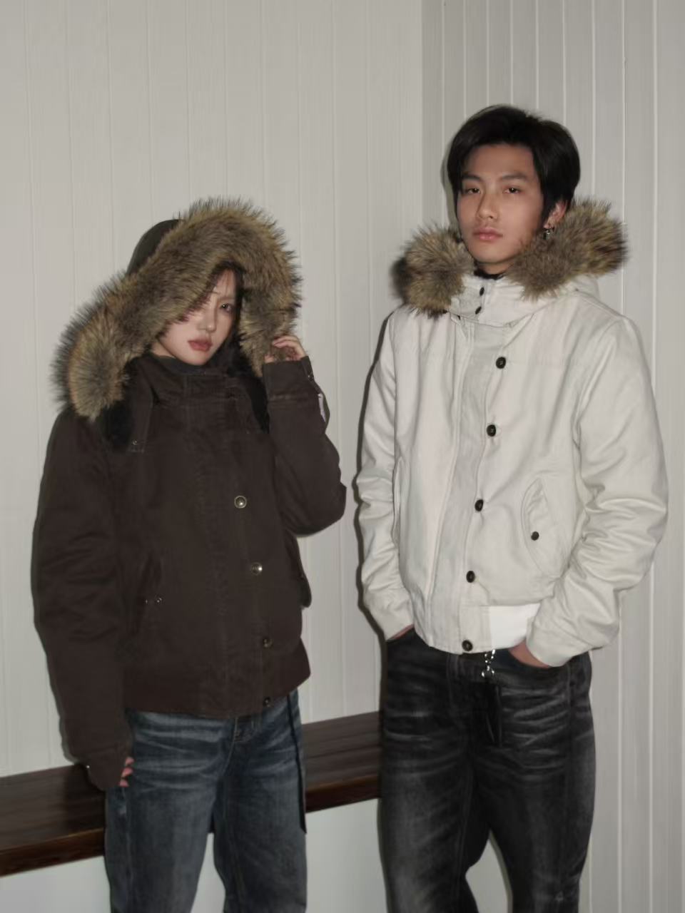 Couple wearing short hooded jackets with fur trimmed hoods, a dark brown one and an off white one, against a white wall