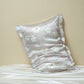 Taupe silk pillowcase printed with pale outline flowers, leaning on a beige wall above white silk