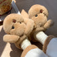 Brown plush slippers with a teddy bear face and short arms worn with cream socks on a wooden floor
