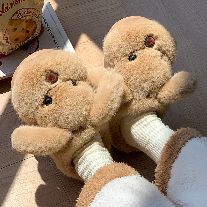 Brown plush slippers with a teddy bear face and short arms worn with cream socks on a wooden floor