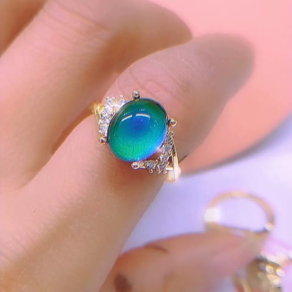 Close-up of a single gold-tone ring with a teal and green color-changing oval cabochon on a fingertip