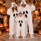 Two adults and a child in matching white plush ghost onesies in a Halloween room with pumpkins and lights