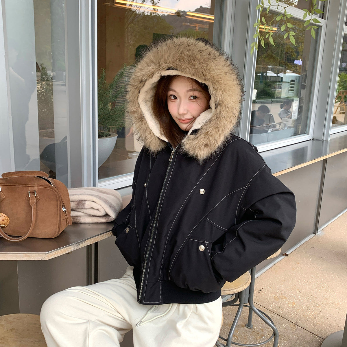 Black hooded bomber jacket with the hood up, vegan fur trim and cream lining, worn seated outdoors
