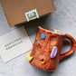 Red orange speckled ceramic mug with blue, lilac and yellow climbing hold decorations beside a box and a card