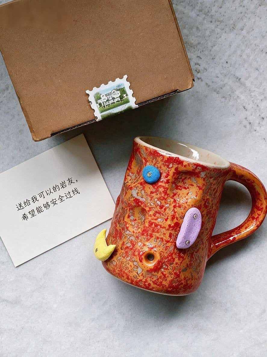 Red orange speckled ceramic mug with blue, lilac and yellow climbing hold decorations beside a box and a card