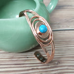 Copper cuff bracelet, blue stone in three concentric ovals, propped on a green ceramic pot on wood