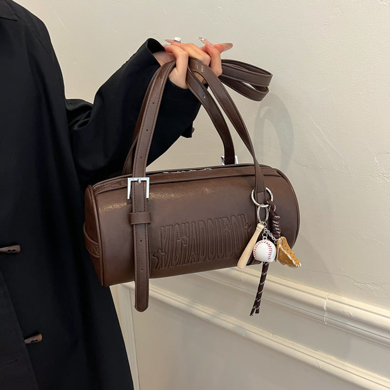 Hand holding a dark brown cylinder shoulder bag with a silver tone buckle strap and a baseball glove charm