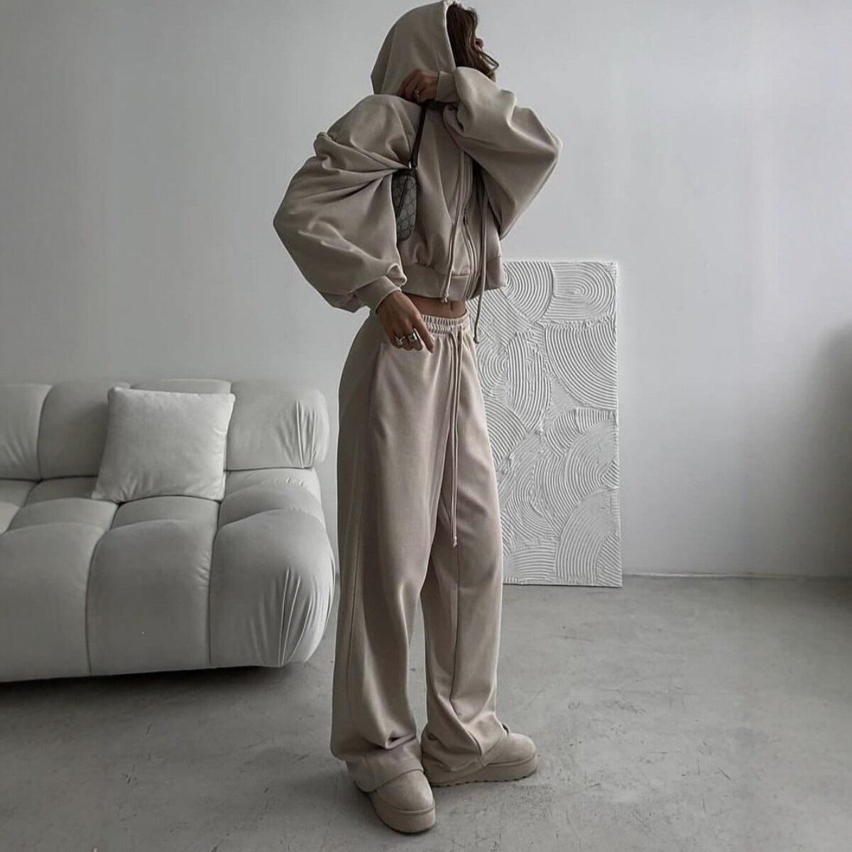 Woman in a taupe khaki hoodie set with the hood up and wide leg trousers, standing beside a sofa