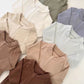 Pile of zip front baby rompers in chocolate, cream, white, olive, lavender gray and peach on a white surface