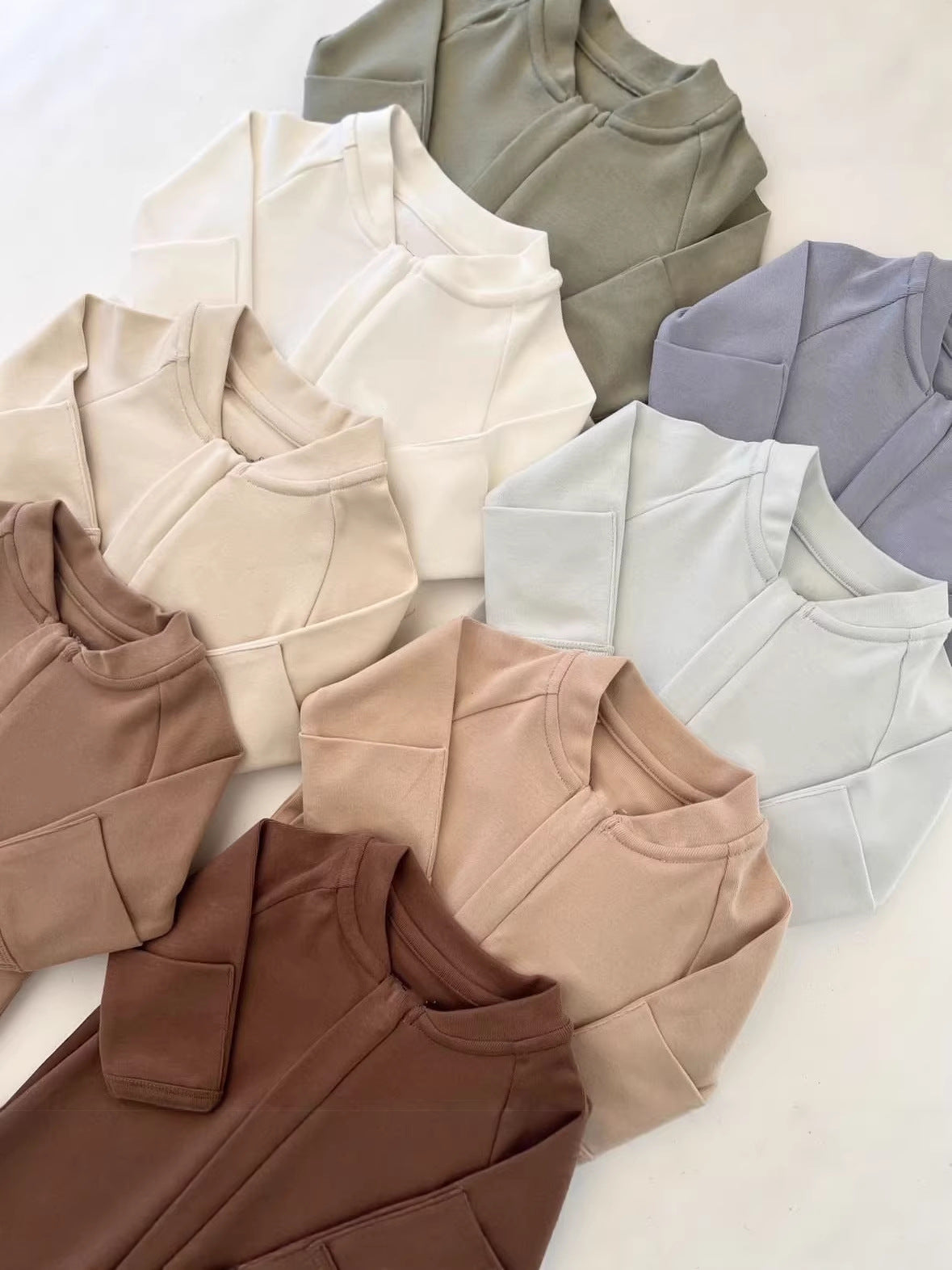 Pile of zip front baby rompers in chocolate, cream, white, olive, lavender gray and peach on a white surface