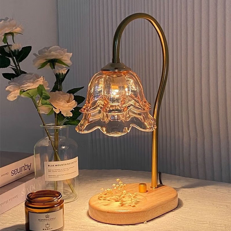 Retro candle warmer lamp with an amber tulip shade lit on a bedside table beside flowers and a candle jar