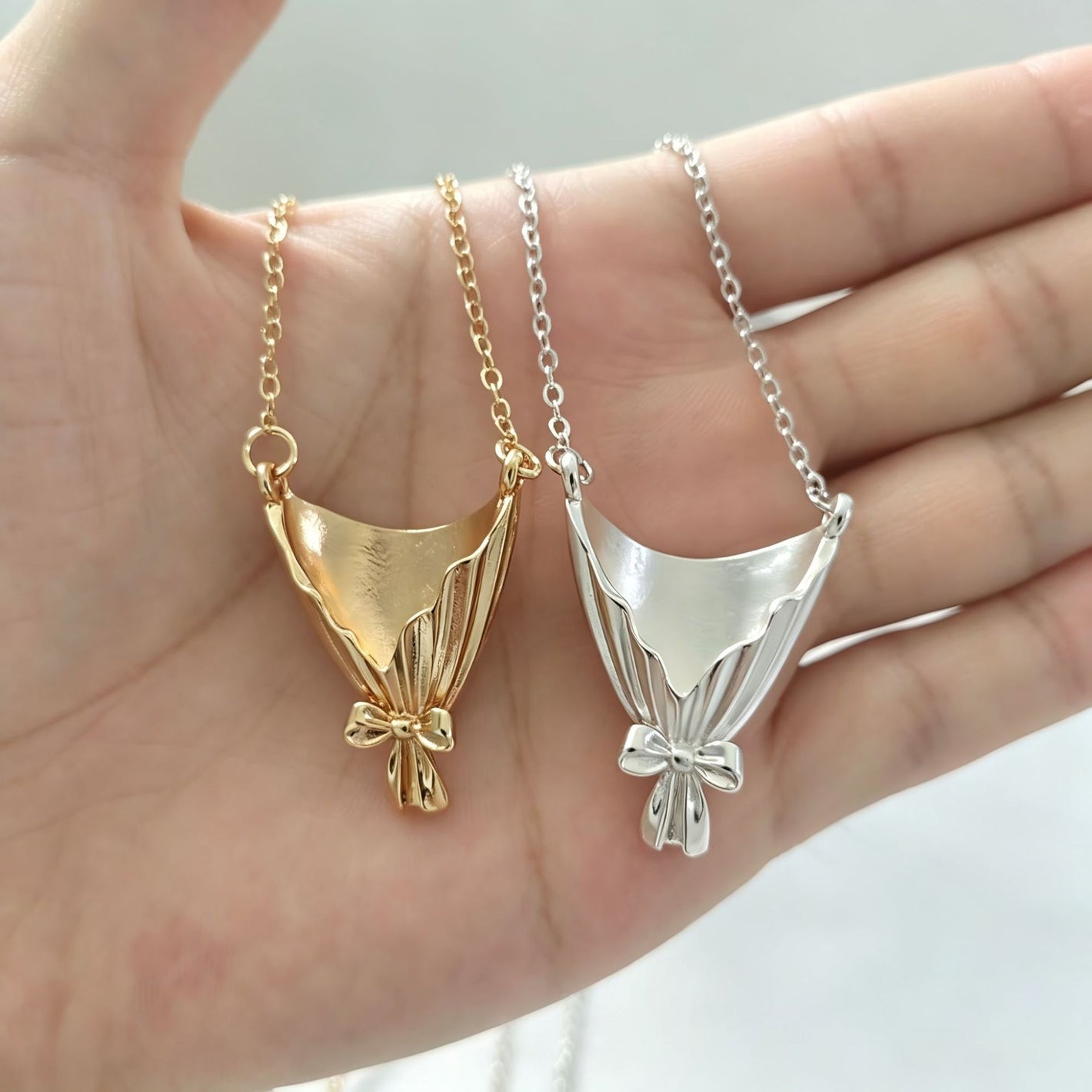Gold tone and silver tone bouquet wrap pendants side by side in a palm, ribbon bows facing forward