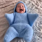 Blue crochet five-point star hooded baby bunting worn by a smiling baby on a bed