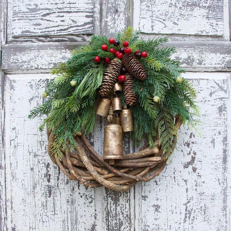 Christmas wreath hanging on a weathered white door, with a rattan ring, pine cones, red berries and gold-tone bells