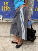 Woman in gray wide-leg pants with white side stripes, a blue tank top and a black bag against a poster wall