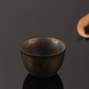 Dark ceramic tea cup with gold fleck glaze on a black reflective surface beside a scoop of loose tea