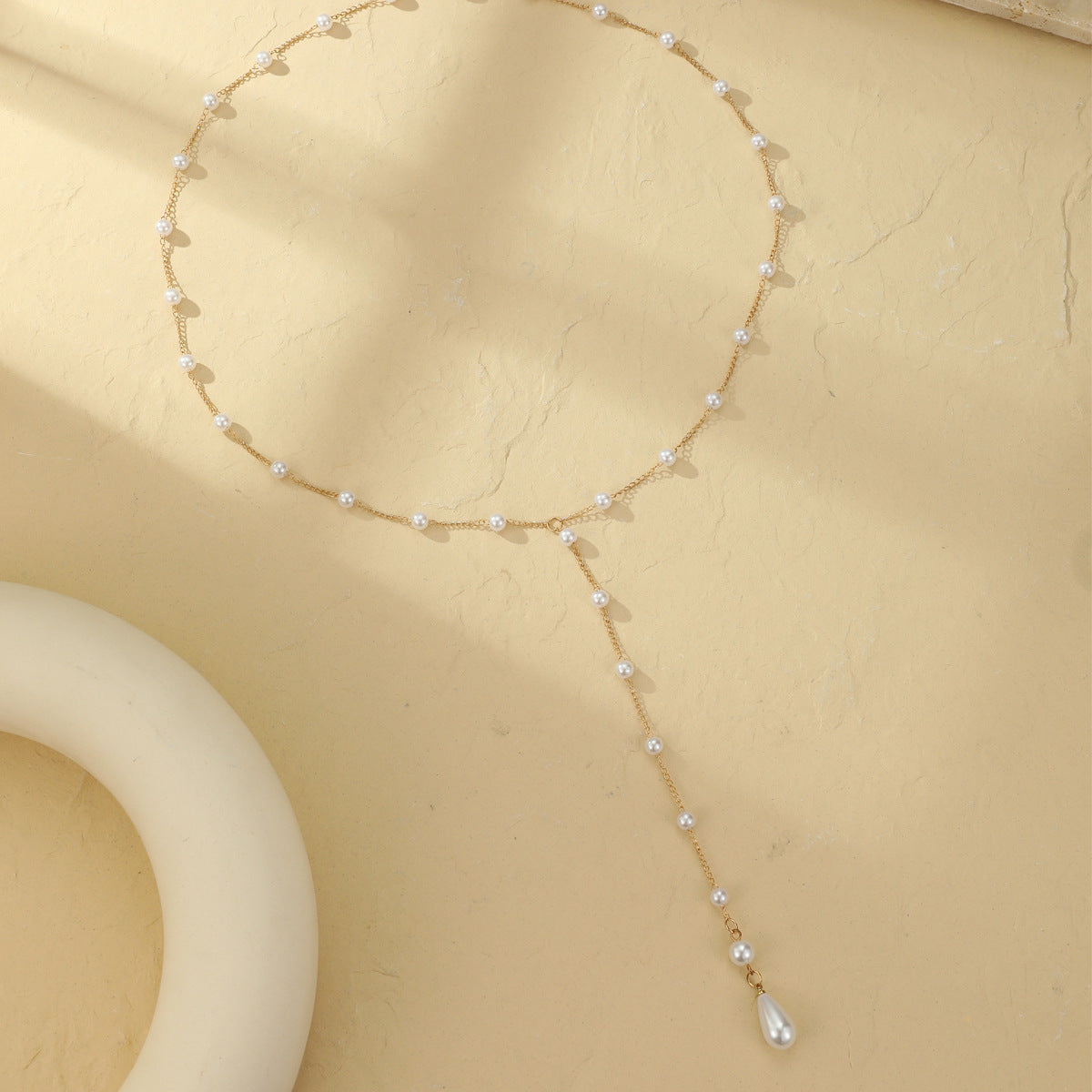 Flatlay of gold tone faux pearl backdrop lariat necklace with round pearls and a teardrop drop on cream